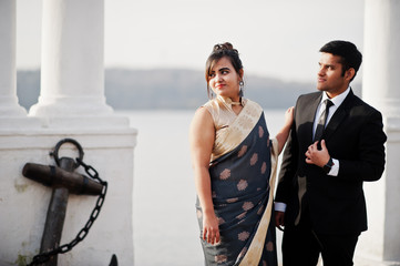 Wall Mural - Elegant and fashionable indian friends couple of woman in saree and man in suit posed on the shore of the marina.