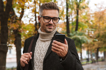photo of masculine man 30s wearing warm clothes walking outdoor through autumn park, and using mobil