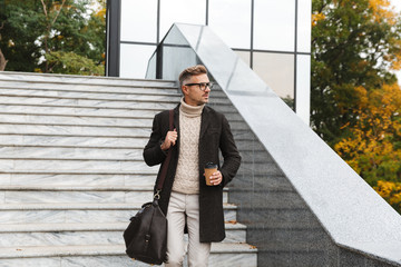 Canvas Print - Image of smart man 30s wearing eyeglasses, walking through city street and holding takeaway coffee