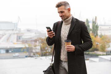 Canvas Print - Portrait of bearded man 30s wearing jacket using mobile phone and holding takeaway coffee, while walking through city street