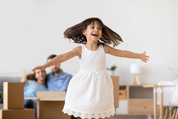 Canvas Print - Excited cute child feel carefree having fun relocating into new home, funny little girl playing jumping enjoying move in big modern living room with parents, happy kid daughter on moving day concept