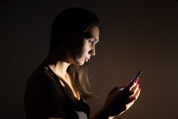 Teen girl excessively sitting at the phone at home. he is a victim of online social networks. Sad teen checking phone sitting on the floor in the living room at home with a dark background