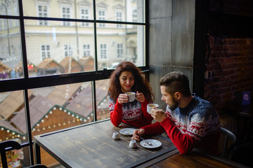 Wall Mural - Happy and romantic couple in warm sweaters drink coffee from disposable paper cups in cafeteria. Holidays, christmas, winter, love, hot drinks, people concept