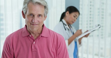 Wall Mural - Close up of senior patient smiling at camera while doctor works in background