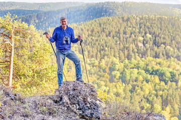 A man with Scandinavian sticks stands on top of a mountain and admires the view of the Ural taiga.