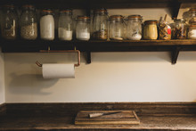 Wooden Chopping Board And Knife On Vintage Wooden Kitchen Cupboard, Row Of Glass Jars With Cooking Ingredients On A Wooden Shelf.