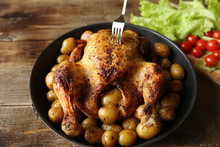 Roasted Chicken With A Golden Crust And Golden Potatoes In A Frying Pan With A Fork In The Chicken, Decorated With Green Salad And Red Tomatoes On A Wooden Rustic Background