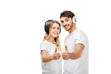 Wall Mural - happy young couple in headphones showing thumbs up and smiling at camera isolated on white