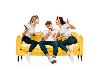 Wall Mural - smiling young women talking and man eating popcorn while sitting together on sofa isolated on white