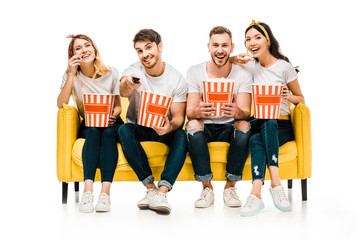  happy young friends holding popcorn boxes and watching tv while sitting on yellow sofa isolated on white