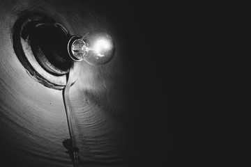 Incandescent lamp burns inside dark room close-up. Illumination. Old light bulb on rough wall. Lamp illuminate basement in grayscale. Light in darkness. Minimalist monochrome background. Copy space.