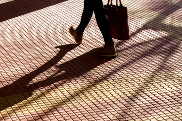 Wall Mural - Blurry silhouette shadow of legs of a person carrying a bag while walking on tiled street sidewalk