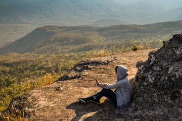 Wall Mural - A woman sitting on rocky mountain using smartphone and looking out at beautiful natural view
