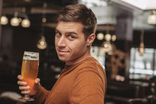 Charming Young Man Smiling To The Camera Over His Shoulder, Having Glass Of Cool Freshly Brewed Craft Beer At The Pub. Attractive Male Customer Relaxing At The Restaurant, Drinking After Work