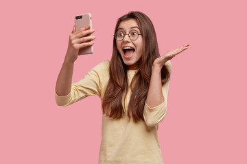 Wall Mural - Happy woman spreads palm, smiles joyfully, holds modern cell phone in front, makes video call, glad to see old friend on distance, isolated over pink background. People, technology, emotions concept