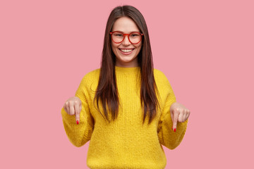 Charming carefree woman with positive expression, points down with both index fingers, dressed in casual clothing, has broad intrested smile, isolated over pink background. Look there, please