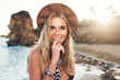 © Look! - Closeup portrait of attractive blonde girl with long hair posing on rocky beach on sunset background. She wears bikini, hat. She holds finger on lips and smiles to the camera.