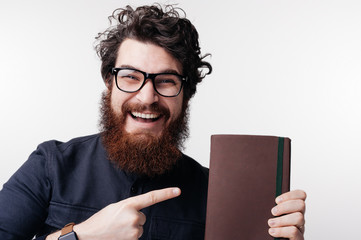 A smiles bearded man in glasses is pointing to his book recommendation, standing over white background