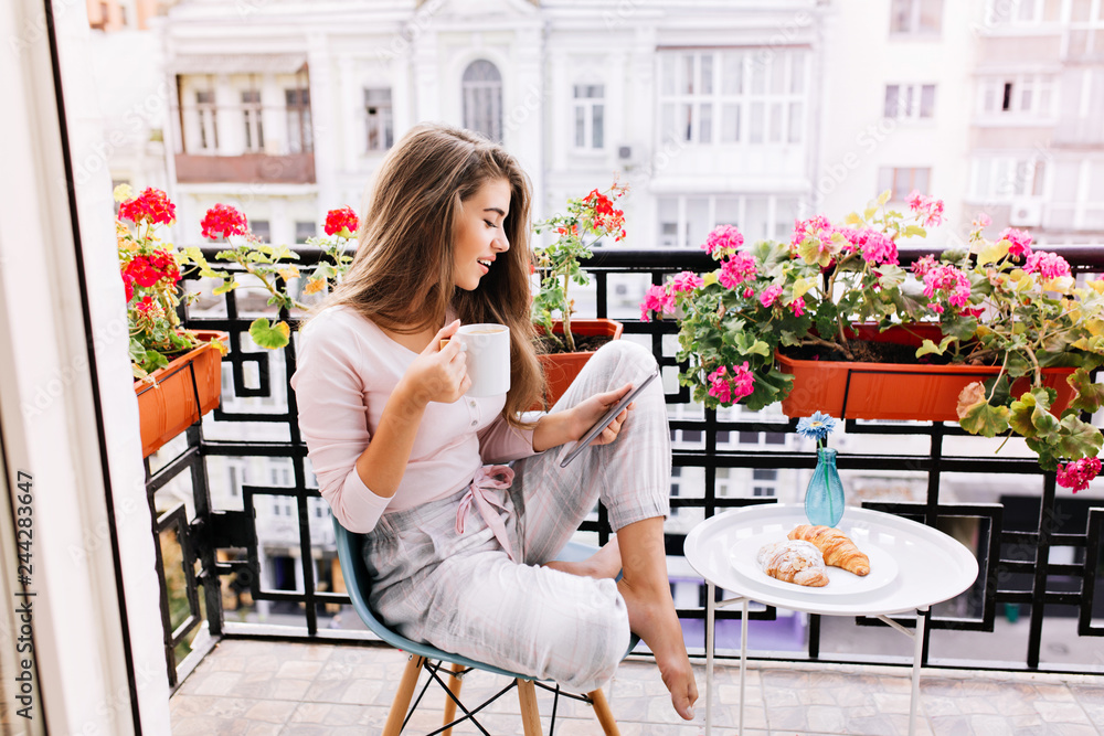 Attractive girl with long hair in pajama having breakfast on balcony in the morning in city. She holds a cup, reading on tablet - obrazy, fototapety, plakaty 