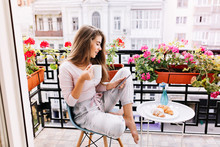 Attractive Girl With Long Hair In Pajama Having Breakfast On Balcony In The Morning In City. She Holds A Cup, Reading On Tablet