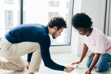 Mixed race couple of students, dressed in casual clothes, sit together on floor in well lit spacious room, prepare for upcoming exams, make project work, compare figures, make notes