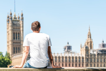 Back of young man with white tshirt sitting on railing in London looking at cityscape skyline of city with Thames River and Westminster Palace during sunny summer