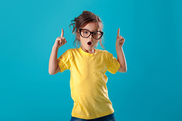 Cute surprised little girl in glasses and yellow t-shirt pointing up