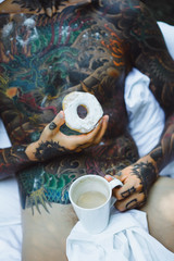 young handsome tattooed man having breakfast in bed in the open air outdoors in the garden, donuts and coffee for breakfast.