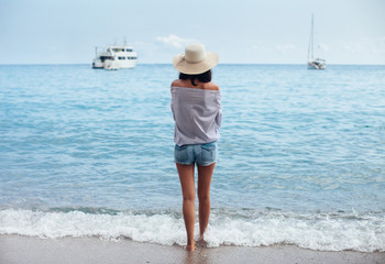 Wall Mural - Back view of young woman in hat enjoy sea and boats from the beach