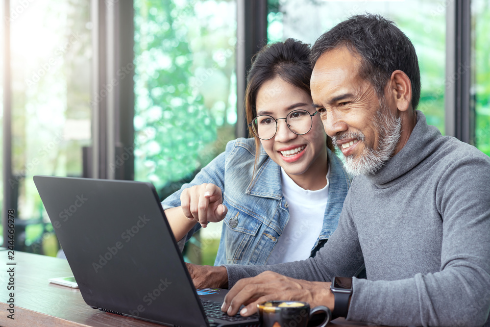 Attractive mature asian man with white stylish short beard looking at laptop computer with teenage eye glasses hipster woman in cafe. Teaching internet online or wifi technology in older man concept. - obrazy, fototapety, plakaty 