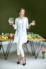 Wall Mural - Full body portrait of a young woman nutritionist in medical gown standing with water in the office with healthy food on the background