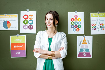 Wall Mural - Portrait of a woman dietitian in medical gown standing on the green wall background with drawings on a topic of healthy food indoors