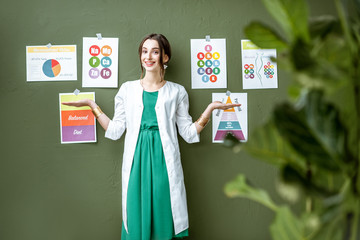 Wall Mural - Portrait of a woman dietitian in medical gown standing on the green wall background with drawings on a topic of healthy food indoors