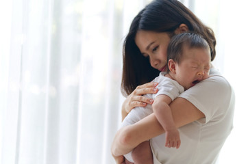 Wall Mural - Cute yawning Asian infant baby on mothers hands standing in the room near the window. Mother hugging baby with love. Happy motherhood and maternity concept.