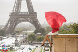 Fototapeta Paryż - Girl hiding herself behind heart shaped red umbrella in front of the Eiffel tower