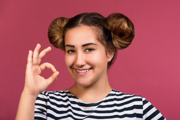 Smiling young hipster girl with playful hairstyle making ok hand gesture isolated over pink background