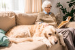 © Seventyfour - Portrait of senior woman reading book and hugging pet dog sitting on couch at home, copy space