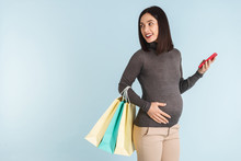Pregnant Woman Isolated Over Blue Wall Background Using Mobile Phone Holding Shopping Bags.