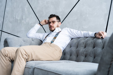 Wall Mural - Enjoying free time. Thoughtful young man in full suit looking away while sitting on the sofa at home.