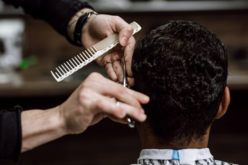 The barber is cutting a man's hair holding scissors and comb in his hands opposite the mirror in a barbershop