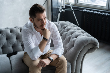 Wall Mural - Enjoying free time. Thoughtful young man in full suit looking away while sitting on the sofa at home.