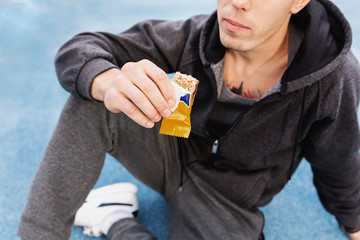 Handsome young guy an athlete in a gray tracksuit bites off with an energy bar after a grueling street workout. Concept of proper nutrition and counting calories