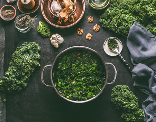 Wall Mural - Stewed kale in cooking pot on rustic kitchen table with ingredients for vegan kale recipes: nuts,garlic, olives oil, top view. Healthy meal.  Detox vegetables .  Clean eating and dieting concept.