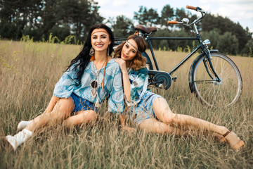 Wall Mural - Two cute beautiful hippie girls outdoors ride a bike, have fun, laugh, dressed in denim shorts, shirts, accessories, bracelets, necklaces, earrings, curly hair, bohemia, indie, boho style, summer