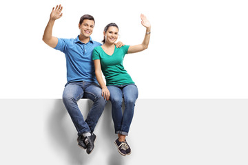 Full length portrait of a young casual couple sitting on a panel and waving at the camera