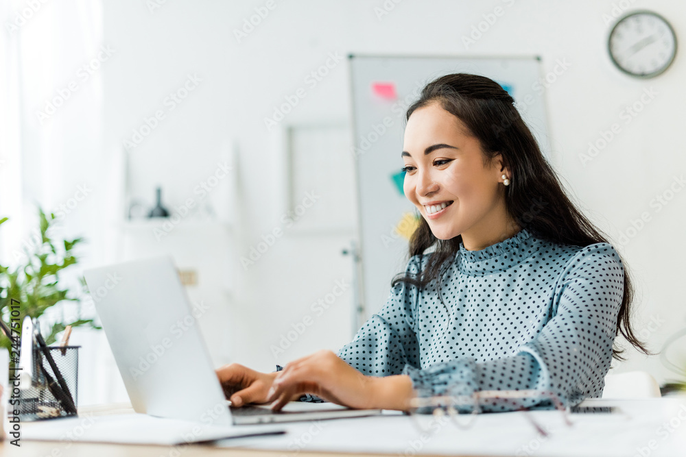 beautiful smiling asian businesswoman sitting at desk and using laptop in office - obrazy, fototapety, plakaty 