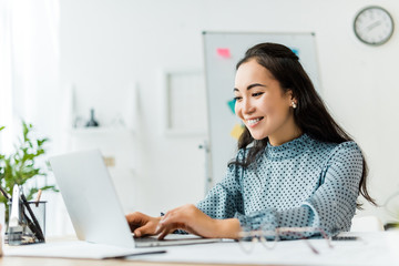 beautiful smiling asian businesswoman sitting at desk and using laptop in office