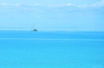 A distant, solitary, traditional, cutter rigged sailboat sitting at anchor in the Bahamas on a calm Caribbean blue sea that stretches out into a blue cloud sky with no visible horizon. Room for copy.