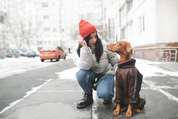 Wall Mural - Attractive woman in warm clothes sitting on the street with a dog looking at a pet and smiling against the backdrop of winter streets and buildings. Winter walk with a dog in doggy clothes.