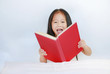 © zilvergolf - Smiling little Asian kid girl reading hardcover book lying with pillow on bed against white background.